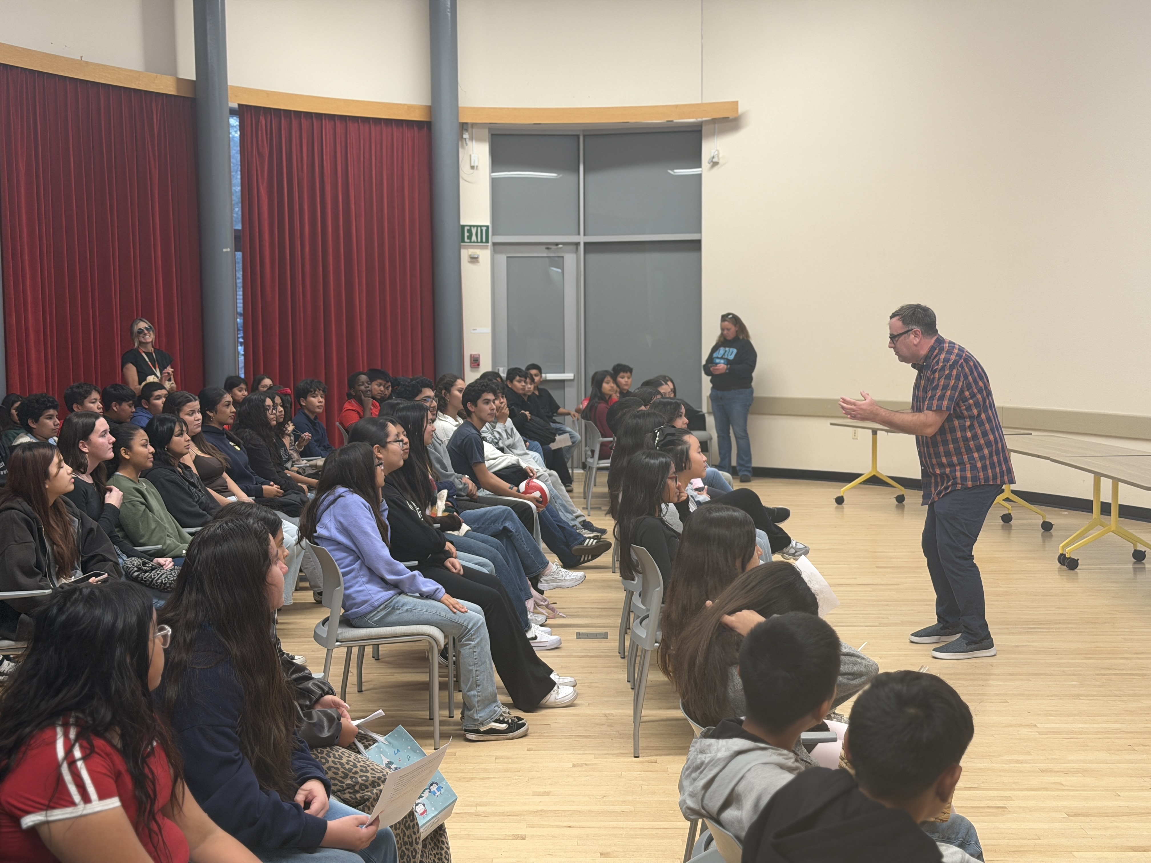 A man wearing a plaid shirt and glasses stands in profile, gesturing with both hands as he addresses a large group of diverse students seated in rows. The room features high ceilings and tall red curtains in the background.