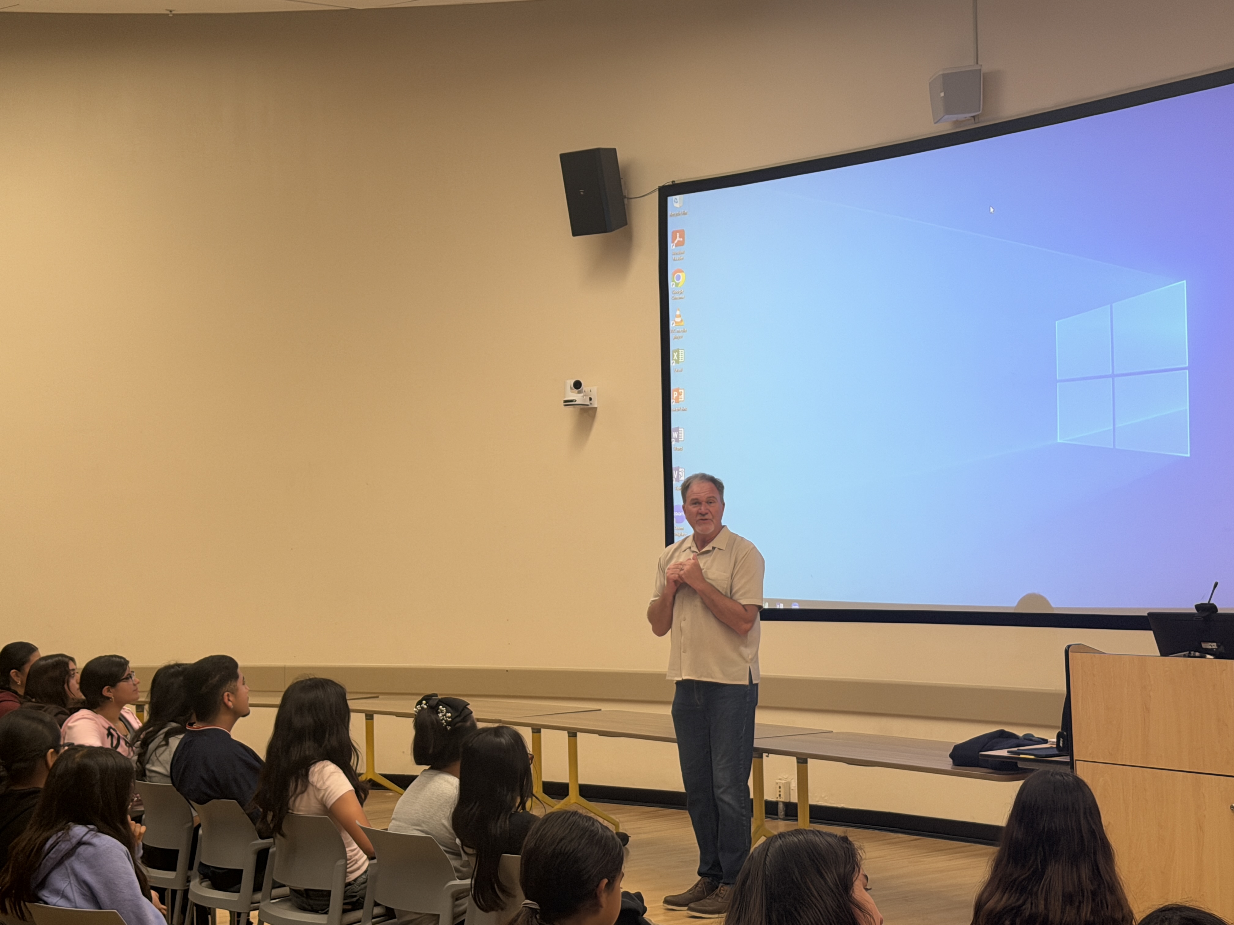 A man in a beige button-down shirt and jeans stands at the front of a classroom, speaking with his hands clasped. To his left, a group of students watches him. Behind him, a large projection screen displays the default blue Windows 10 desktop background.