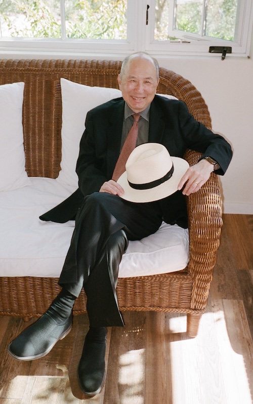 Alan Liu, distinguished professor of English seated on a wicker sofa with white cushions, wearing a dark suit and brown tie, holding a white hat with a black band. The background includes windows with visible greenery.