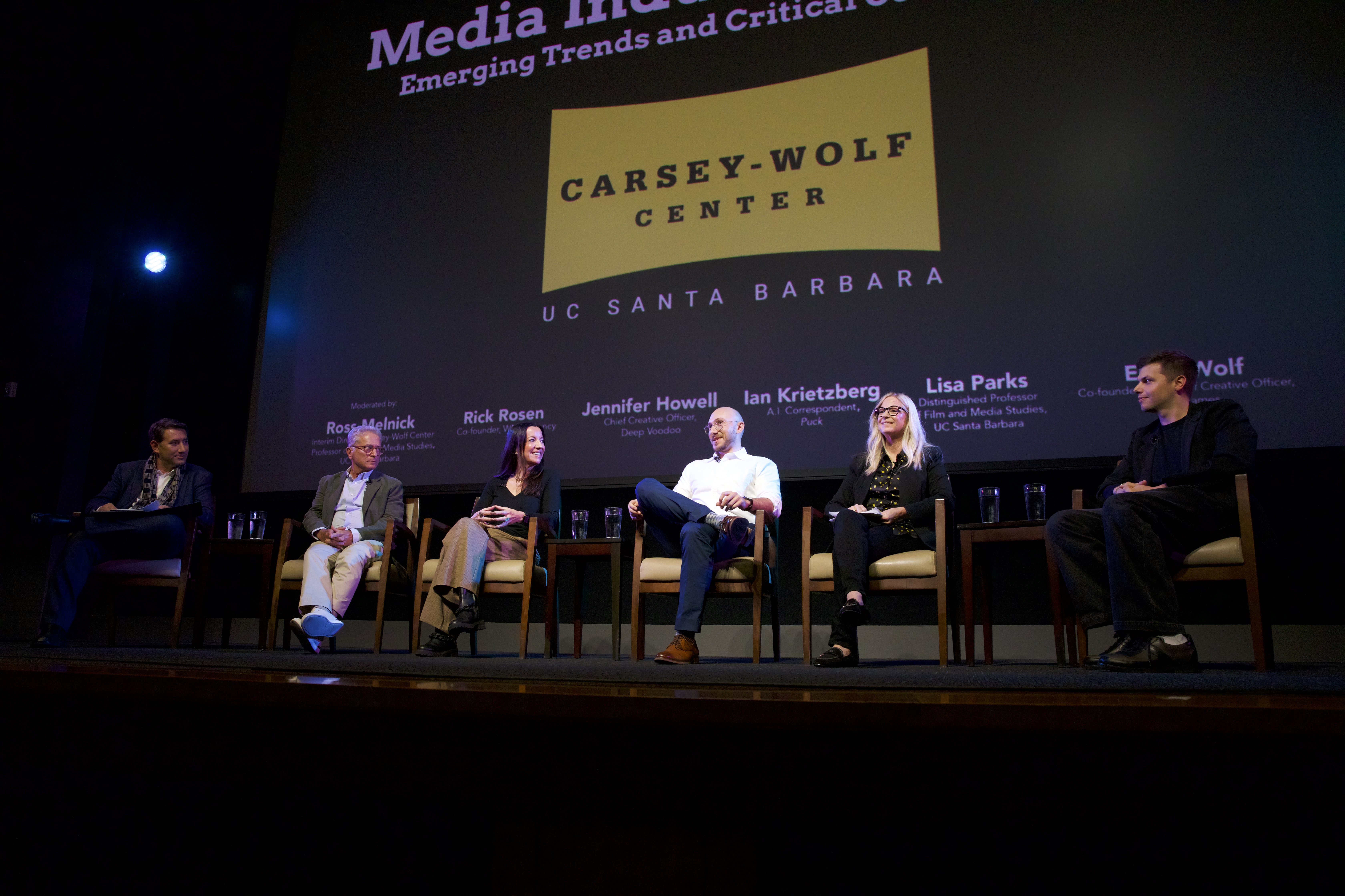 A photograph of a panel discussion on a dimly lit stage. Six people are seated in a row facing the audience. Behind them, a large projection screen reads 'CARSEY-WOLF CENTER' and 'UC SANTA BARBARA'. Below the logos, a list of participants is visible, including 'Moderated by Ross Melnick,' 'Rick Rosen,' 'Jennifer Howell,' 'Ian Kretzberg,' 'Lisa Parks,' and a final panelist named Wolf.