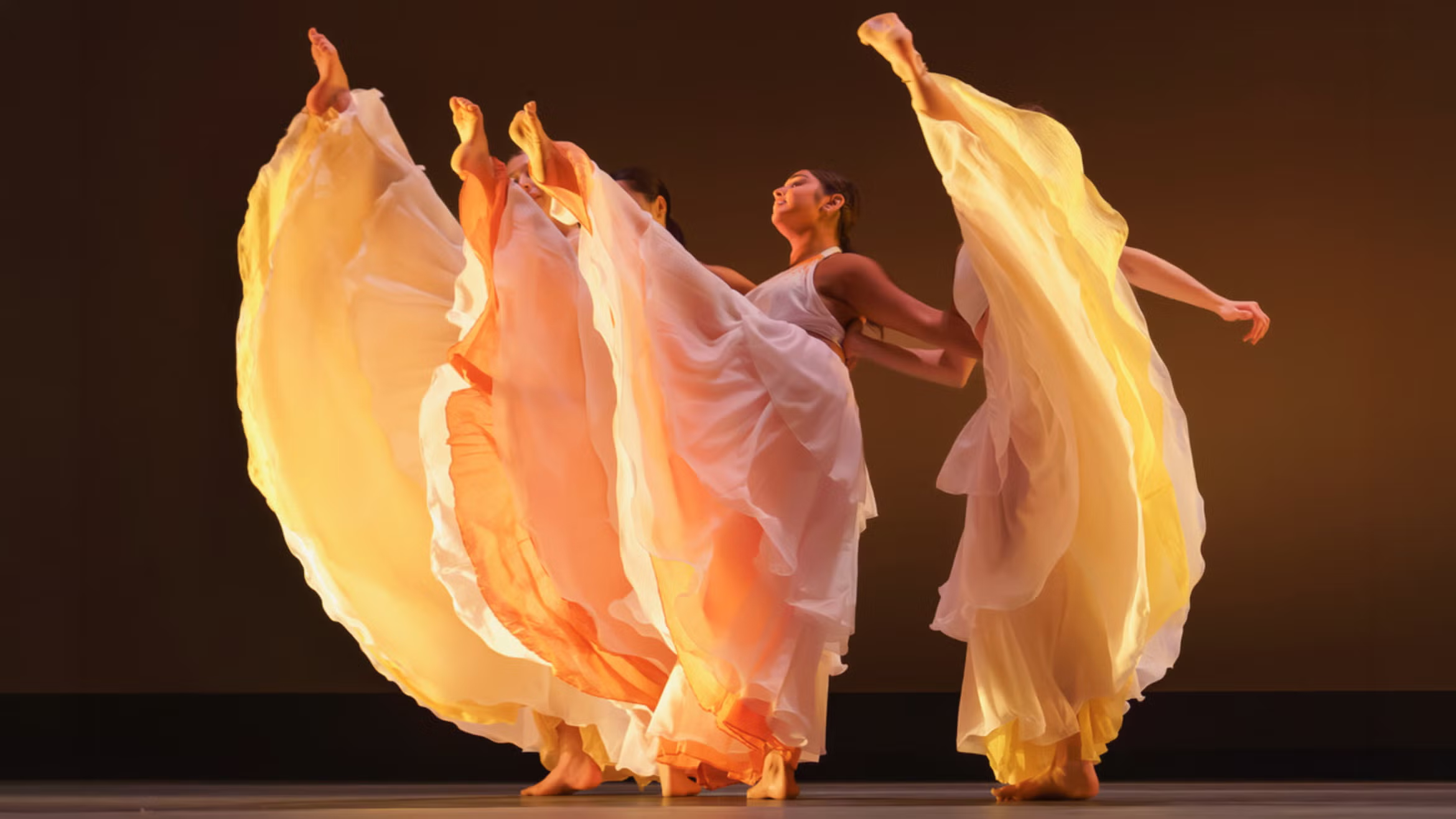 Four dancers with flowy yellow and white skirts performing a high leg extension on stage under warm lighting.
