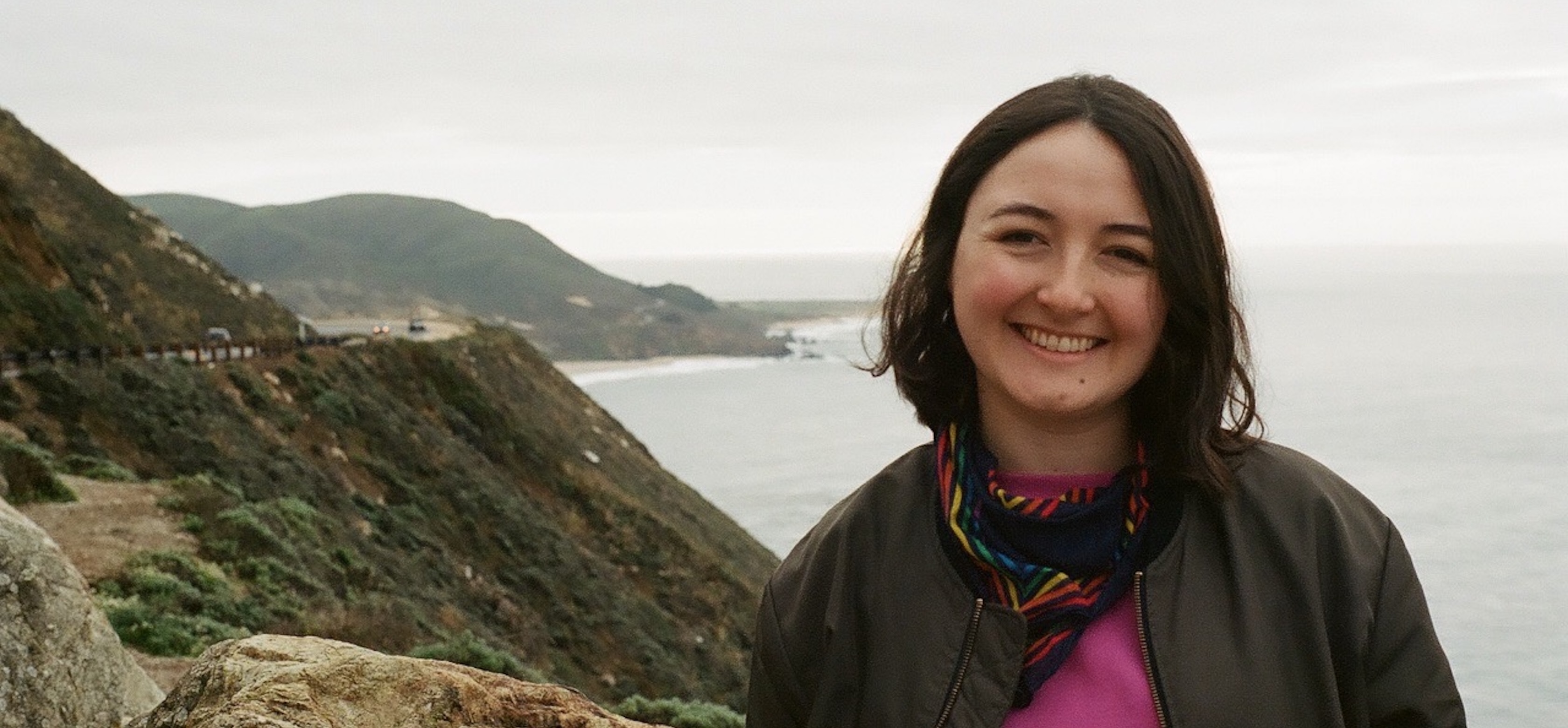 Headshot of Alida Jekabson smiling outdoors, wearing a dark jacket and colorful patterned scarf, with a rocky coastline and the ocean in the background.