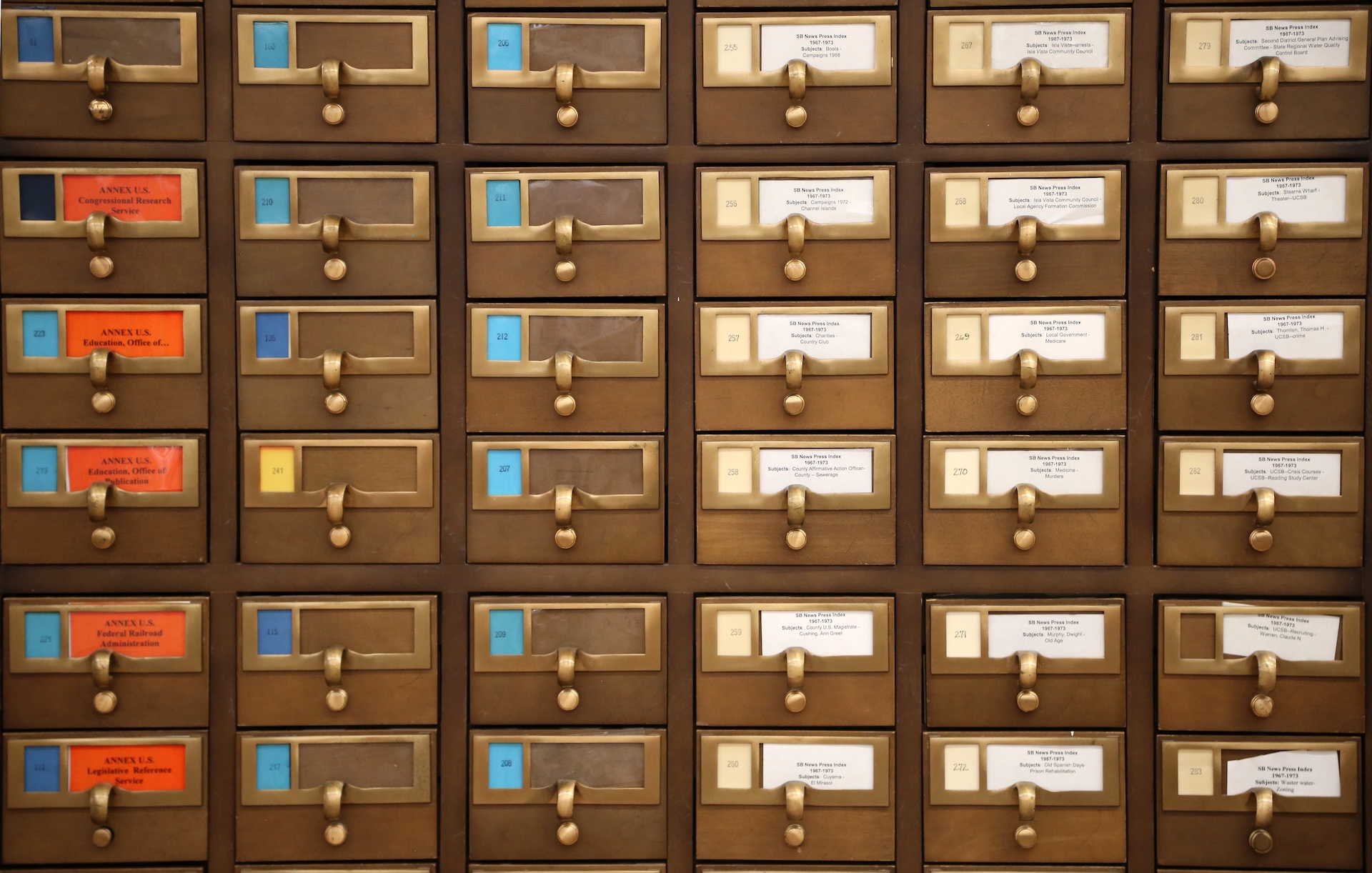 A close-up view of a vintage wooden library card catalog cabinet featuring rows of small drawers with brass handles and printed labels.
