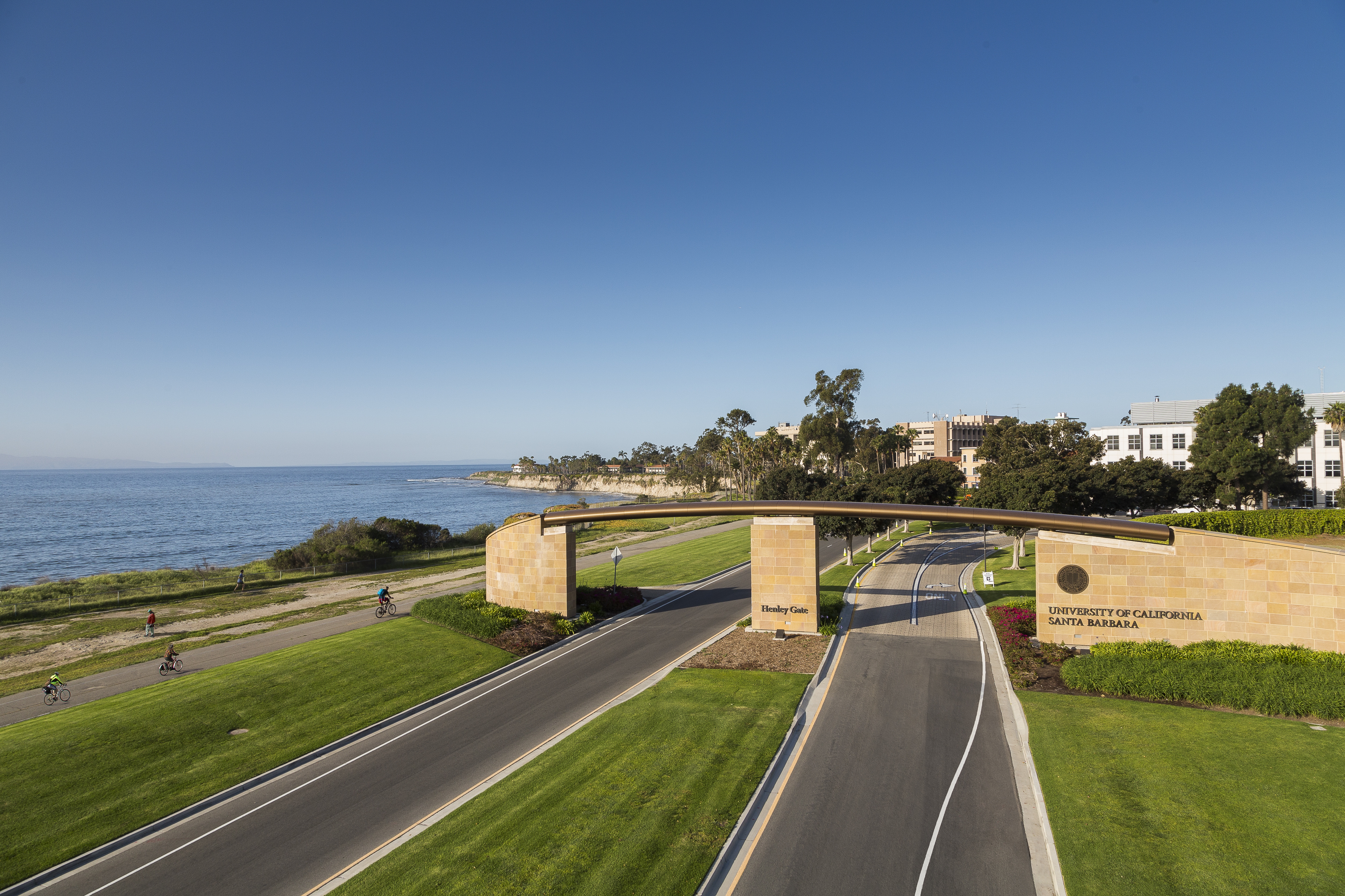 Aerial view of the University of California, Santa Barbara (UCSB) campus entrance at Henley Gate, a large stone archway spanning the divided entrance road. The Pacific Ocean and coastline are visible to the left, with the campus buildings and trees to the right and above the arch. A paved bike path runs along the ocean.