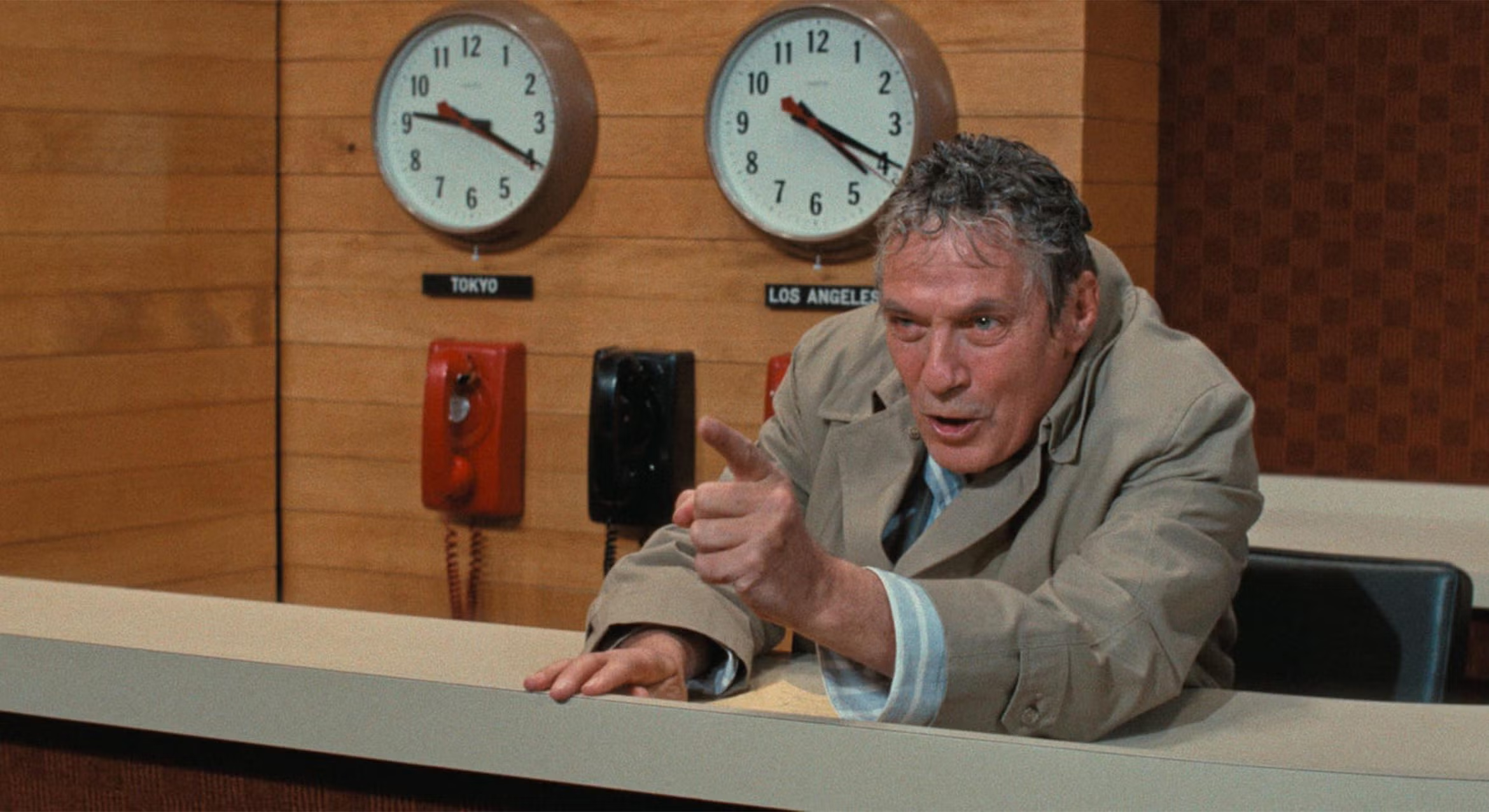Man sitting at a desk with clocks reading Tokyo and Los Angeles in the background points his finger angrily.