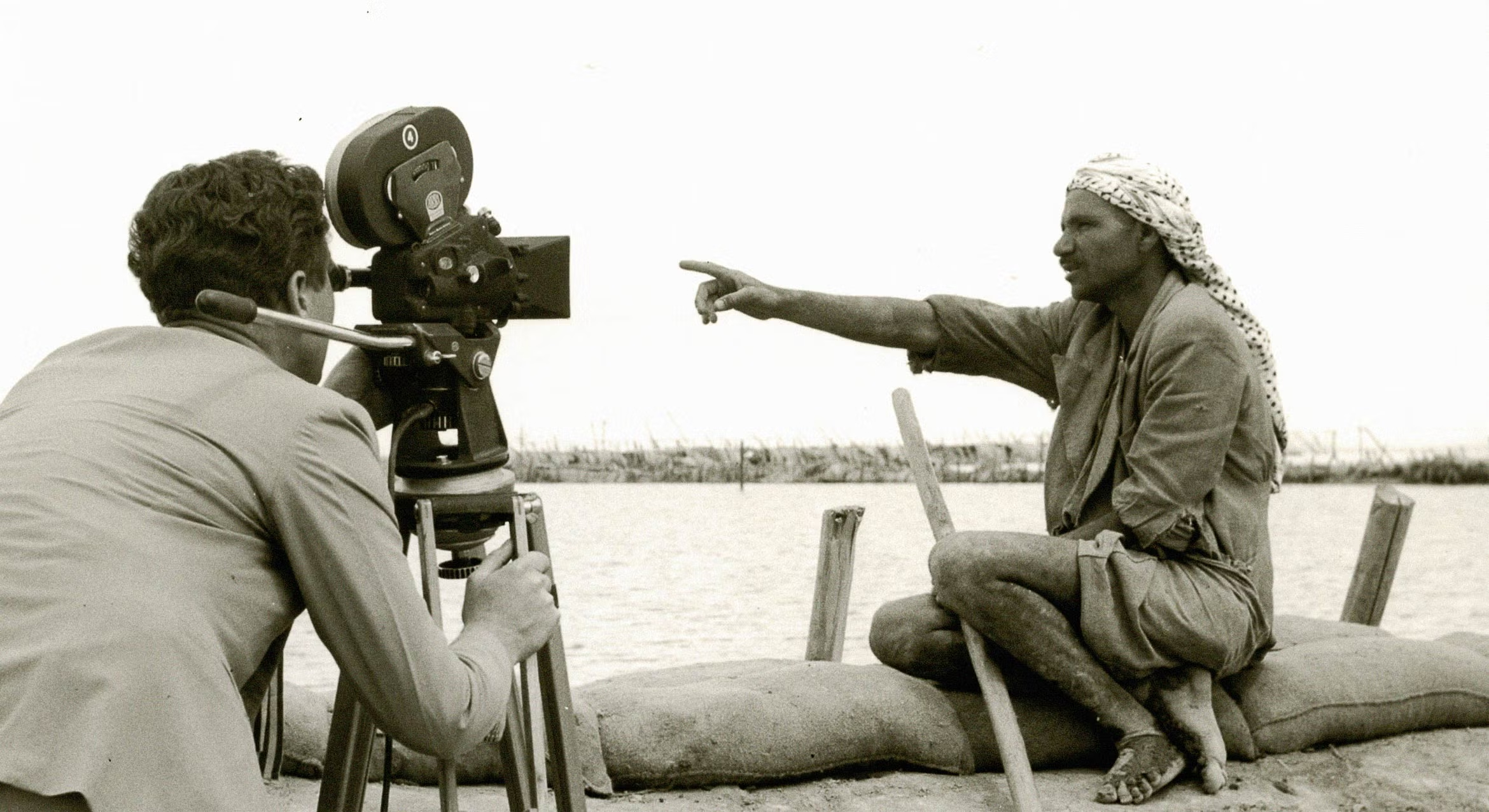 Black and white archival photograph of two men in the desert, with one man seated on sandbags pointing out to the horizon while the other operates a 1950s motion film camera on a tripod.