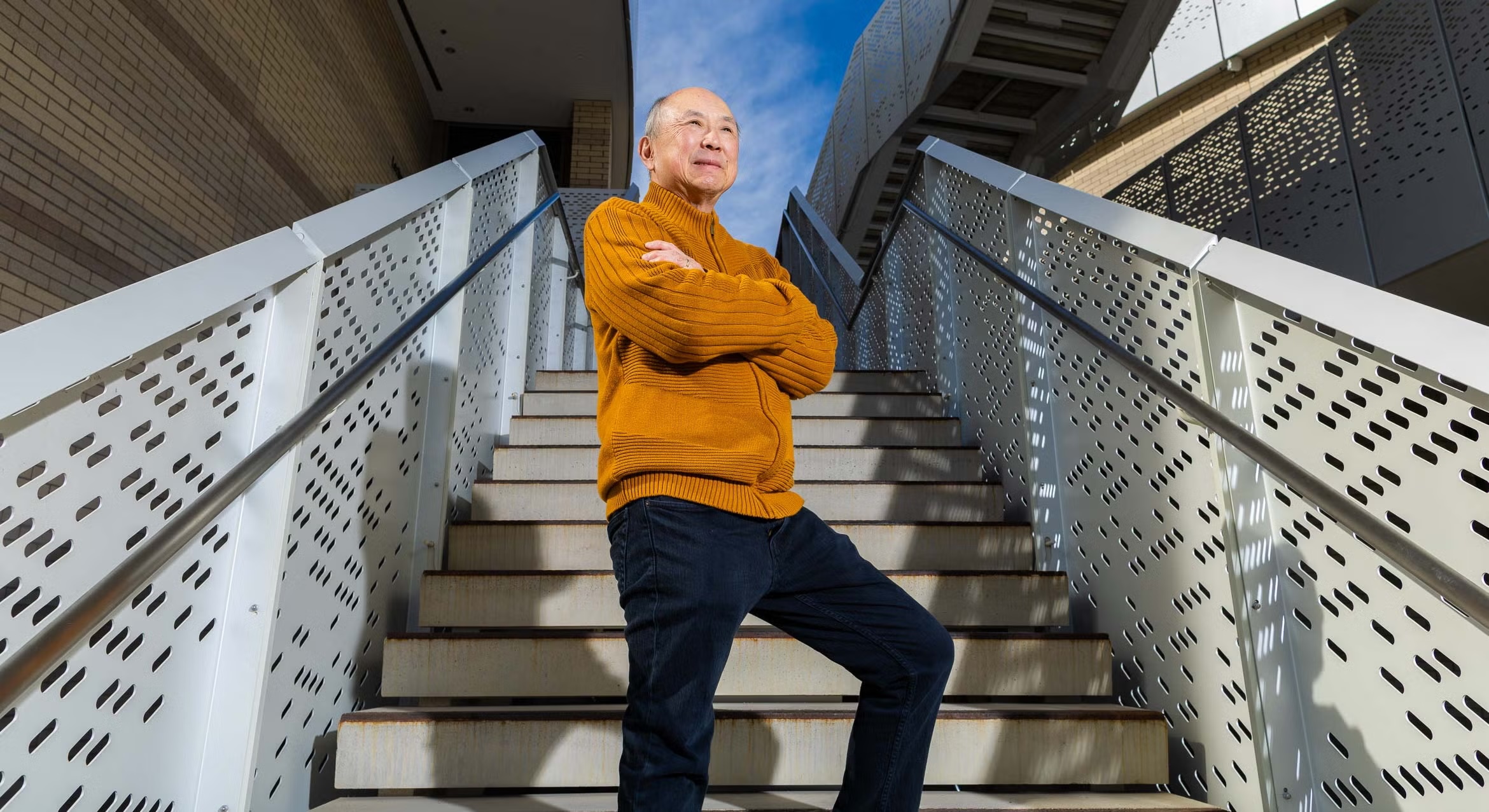 Low-angle portrait of Alan Liu wearing an orange zip-up sweater and jeans, standing on outdoor concrete stairs with his arms crossed, smiling and looking off into the distance against a blue sky.